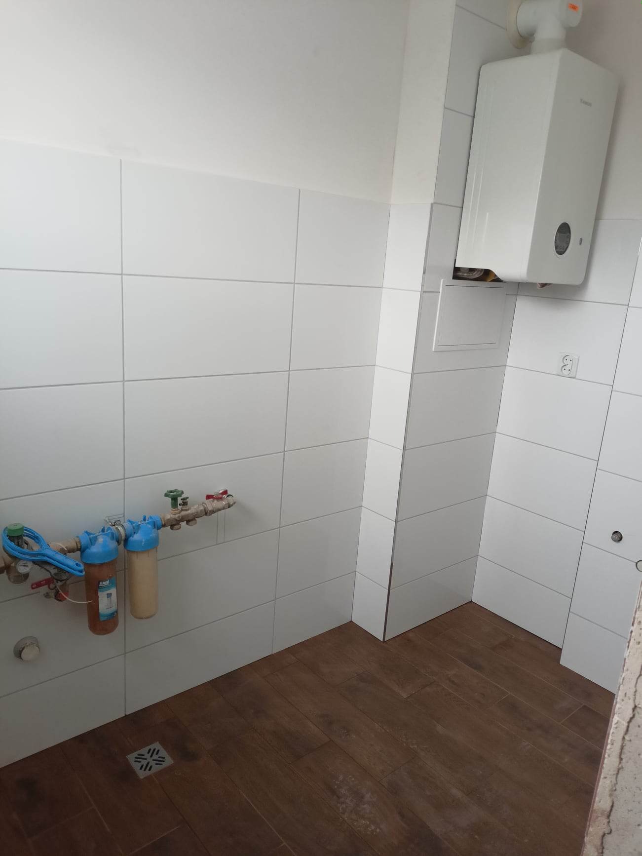 Biała łazienka z brązową podłogą imitującą drewno, widoczne filtry do wody podłączone do rur na ścianie wyłożonej białymi płytkami oraz biały piec gazowy.
