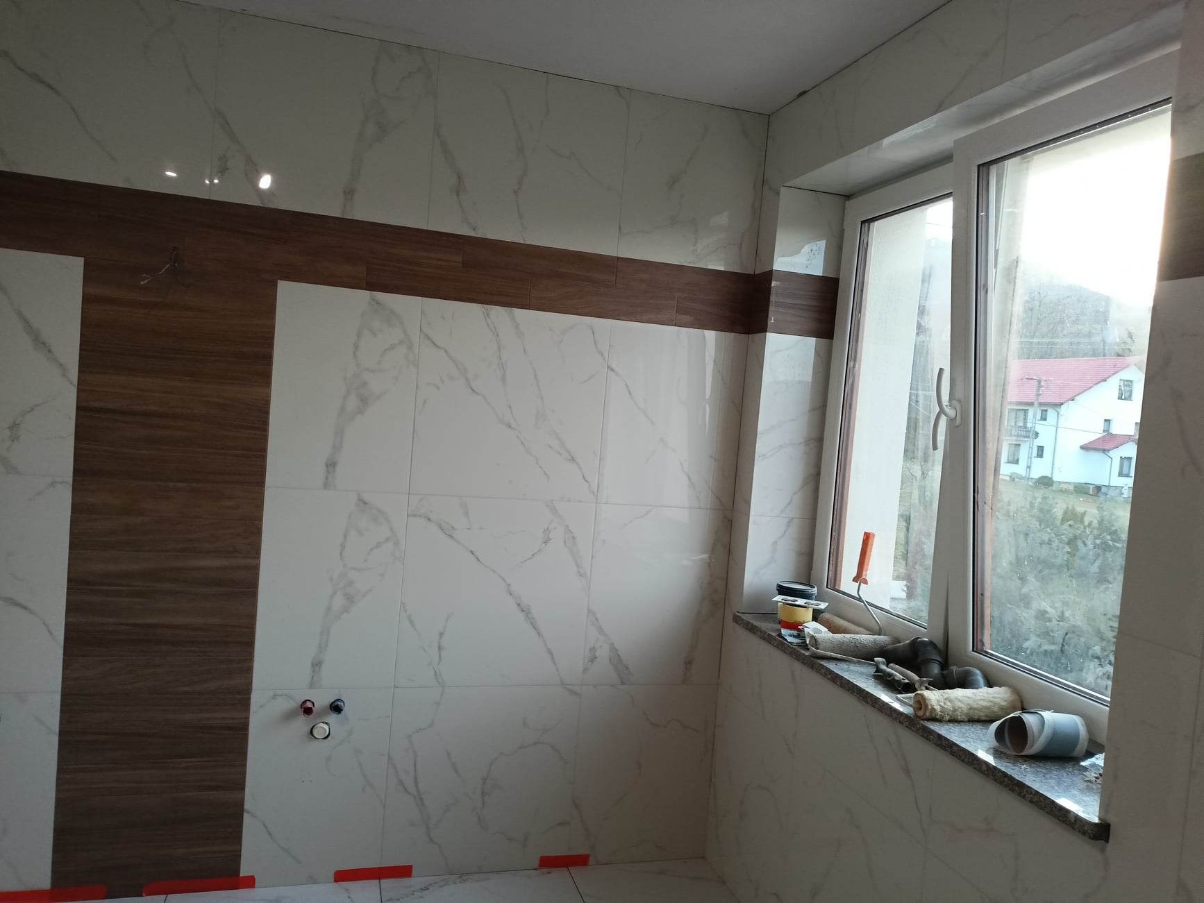 Łazienka w trakcie wykańczania: marmurowe płytki z brązowym pasem dekoracyjnym, widoczne narzędzia malarskie na parapecie, instalacja wodna wystaje ze ściany.