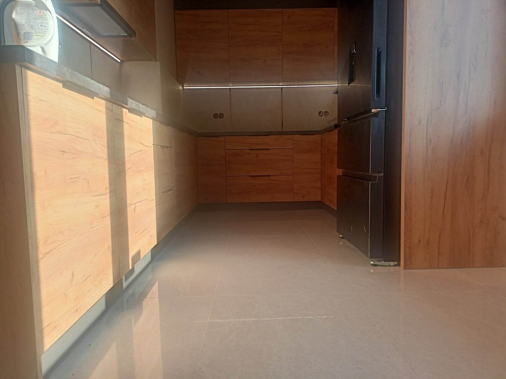 Nowoczesna kuchnia z drewnianymi frontami szafek, szarymi blatami i podświetleniem LED pod szafkami wiszącymi, widoczna lodówka w zabudowie i jasne płytki podłogowe.