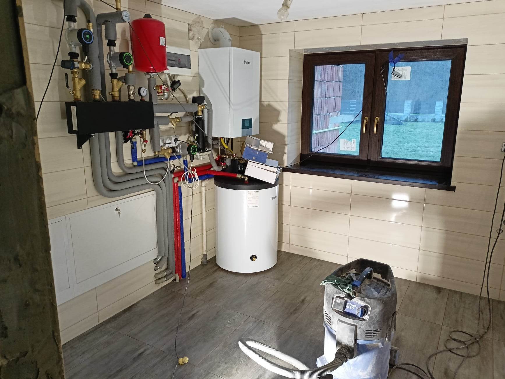 Wnętrze kotłowni z zamontowanym piecem gazowym Vaillant, zasobnikiem ciepłej wody, rozbudowaną instalacją grzewczą z rurami w izolacji, widoczny odkurzacz przemysłowy Nilfisk Alto, ściany wyłożone...