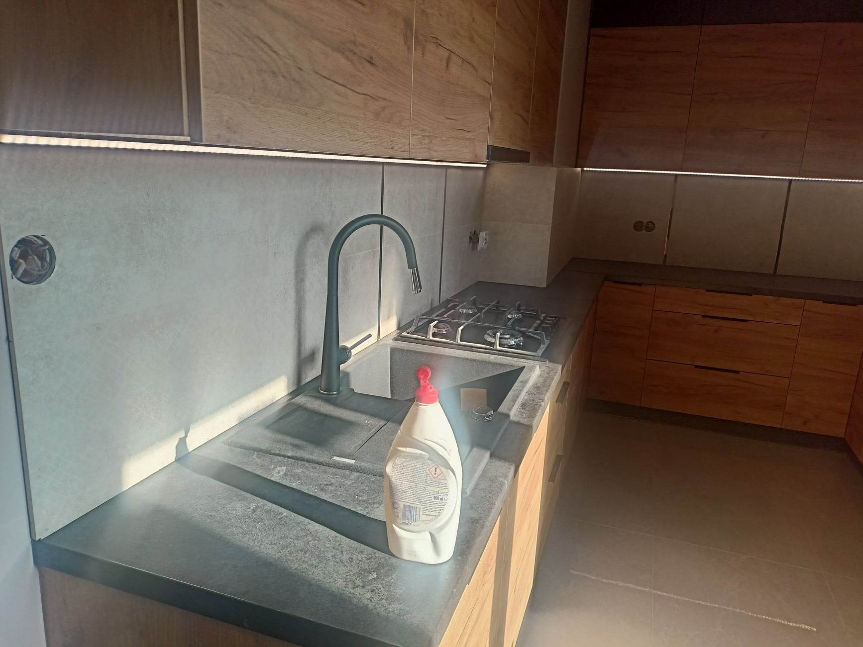 Nowoczesna kuchnia z drewnianymi szafkami, szarą płytą nad blatem i czarną armaturą; butelka płynu do naczyń na blacie.