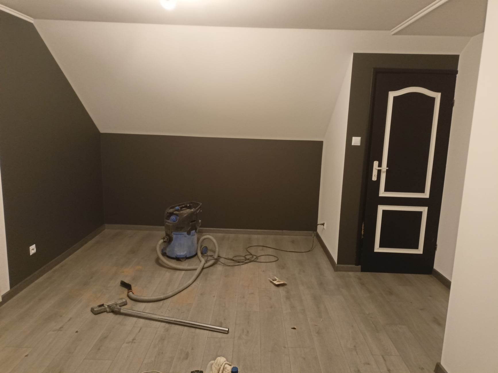 Pokój na poddaszu z pomalowanymi ścianami w kolorze białym i brązowym, widoczny odkurzacz przemysłowy i akcesoria do sprzątania po pracy.