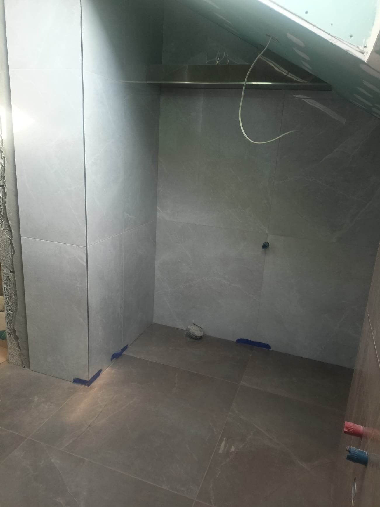 Wnętrze łazienki w trakcie wykańczania, widoczne szare płytki na ścianach i podłodze, fragment sufitu z płyt kartonowo-gipsowych, niebieska taśma malarska zabezpieczająca narożniki oraz wystające...