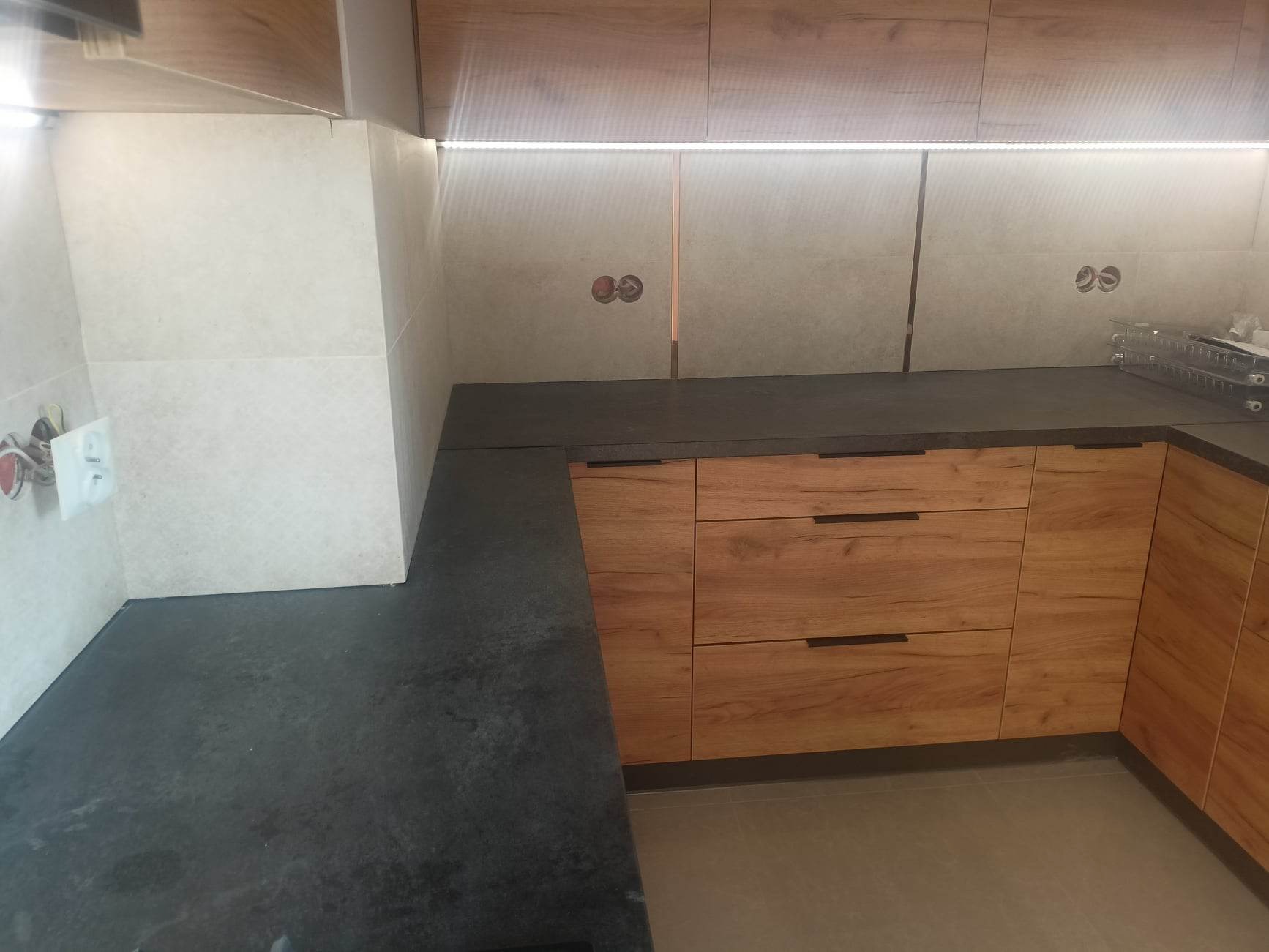 Nowoczesna kuchnia z drewnianymi szafkami, ciemnym blatem i jasnymi płytkami na ścianie, oświetlenie LED pod szafkami górnymi.