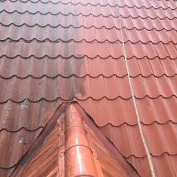 Cleanhouse_warszawa - Częściowo umyty dach z blachodachówki w kolorze ceglastym, widoczna wyraźna różnica między czystą a zabrudzoną powierzchnią, zarys komina w tle.