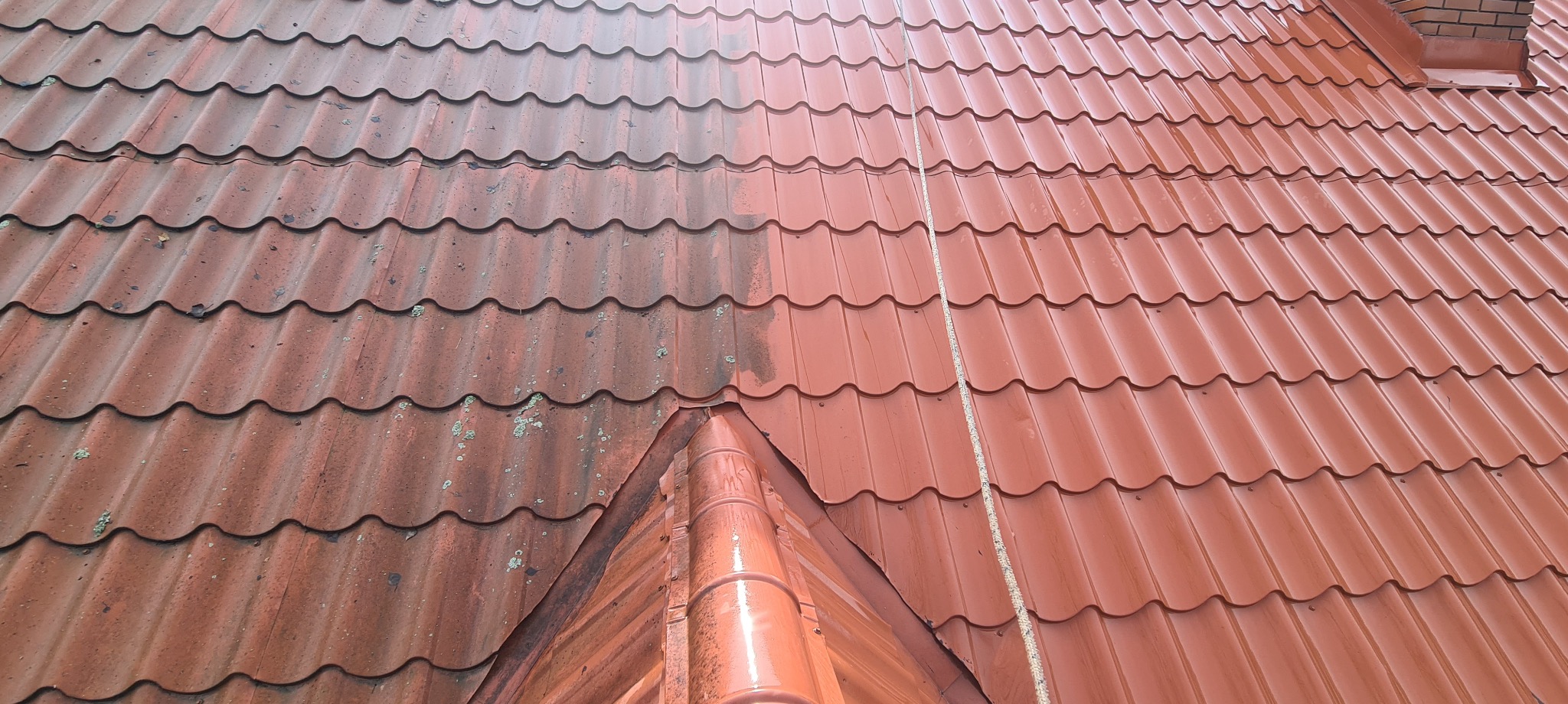 Częściowo umyty dach z blachodachówki w kolorze ceglastym, widoczna wyraźna różnica między czystą a zabrudzoną powierzchnią, zarys komina w tle.