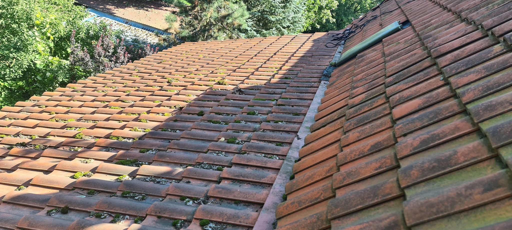 Czerwona dachówka ceramiczna pokryta mchem i porostami, widok z góry, z elementami instalacji na krawędzi dachu oraz zielenią w tle.