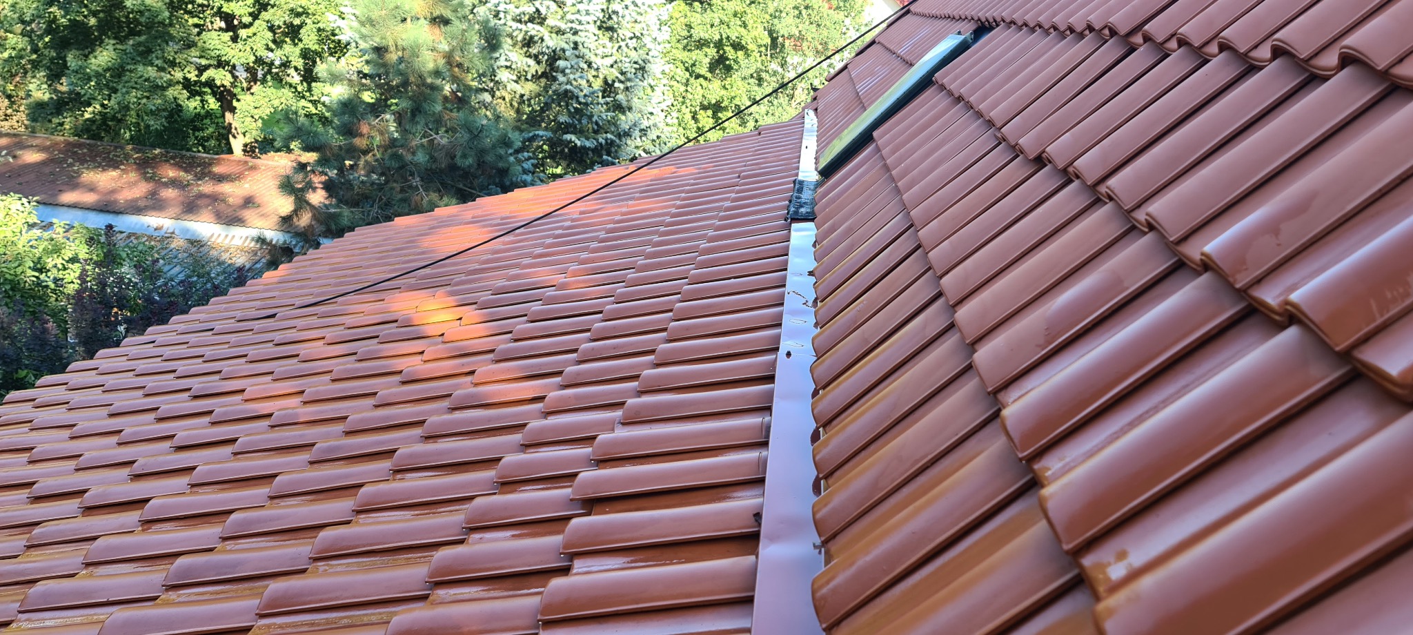 Błyszczące, brązowe dachówki ceramiczne na dachu domu, widok z góry na fragment dachu z zamontowanym oknem dachowym i rynną, w tle zieleń drzew.