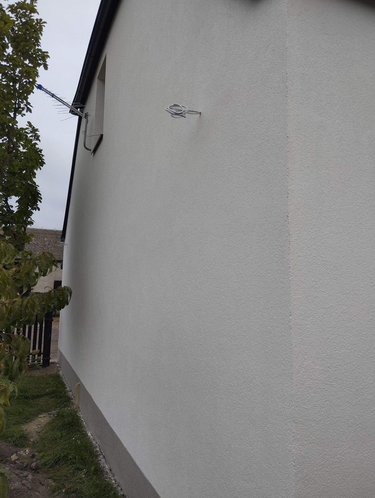 Boczny widok na białą elewację domu z anteną i fragmentem zieleni. Tekstura ściany z bliska, widoczne detale wykończenia. Ujęcie z dołu, pochmurne niebo w tle.