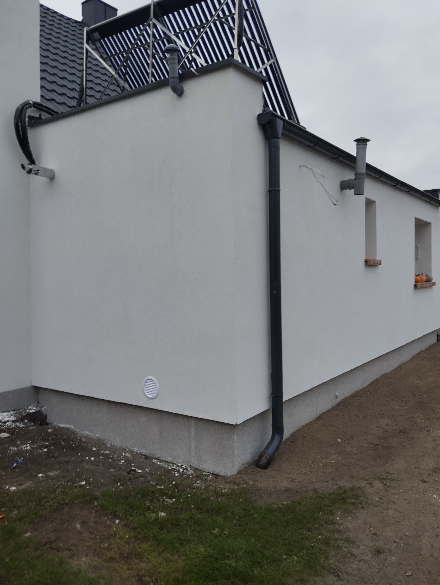 Biała elewacja budynku z szarym dachem i rynnami. Widoczne okna z dekoracjami. Częściowo widoczny system solarny na dachu. Trawa i ziemia wokół budynku.