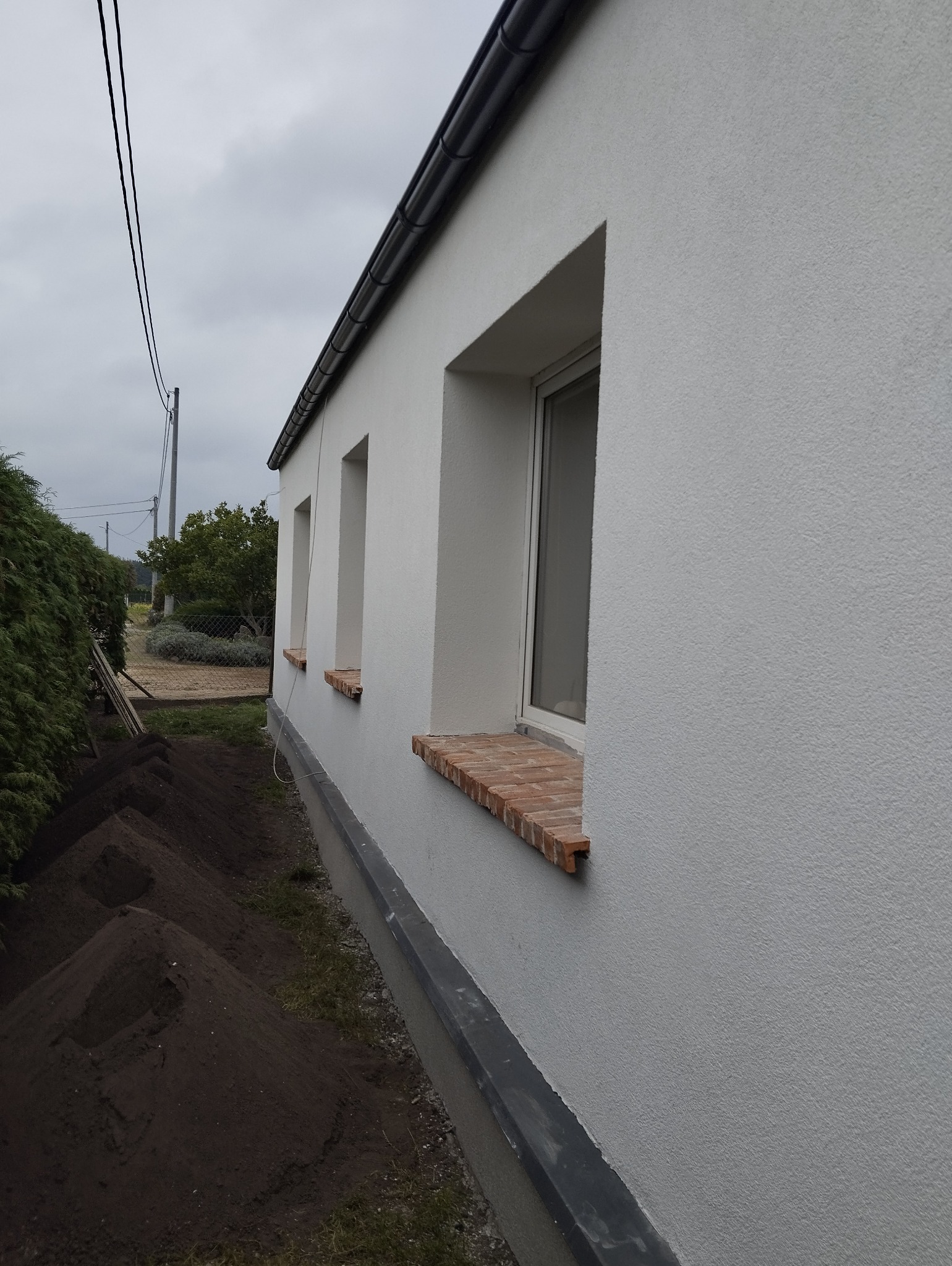 Biała elewacja domu jednorodzinnego z oknami z ceglanymi parapetami, widok z boku, obok wykopany rów z ziemią, pochmurne niebo w tle.