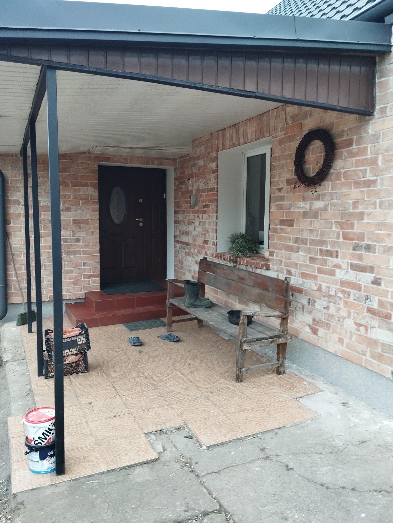 Zadaszone wejście do domu z ceglaną elewacją. Widoczna ławka, doniczka w oknie, wieniec na ścianie i wiadro z farbą. Częściowo położona terakota na podłodze.