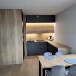 Nowoczesna kuchnia z szarymi szafkami dolnymi i drewnopodobnymi górnymi, oświetleniem podszafkowym LED, drewnianym blatem i stołem z szarymi krzesłami w tle.