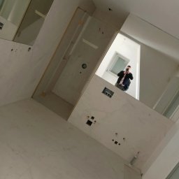 RemBud Hryb - Wnętrze łazienki w trakcie wykańczania, widoczne białe płytki imitujące marmur na ścianach i podłodze, kabina prysznicowa z elementami w kolorze złotym, otwory pod instalacje elektryczne,...