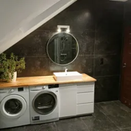 Nowoczesna łazienka na poddaszu z ciemnymi płytkami, drewnianym blatem, białą umywalką nablatową, pralką i suszarką obok siebie, okrągłym lustrem z oświetleniem i rośliną doniczkową.