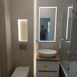 RemBud Hryb - Nowoczesna łazienka z beżowymi płytkami, podświetlanym lustrem i wnęką, umywalką nablatową na drewnianym blacie i wiszącą toaletą; widoczne naklejki na szafce pod umywalką.