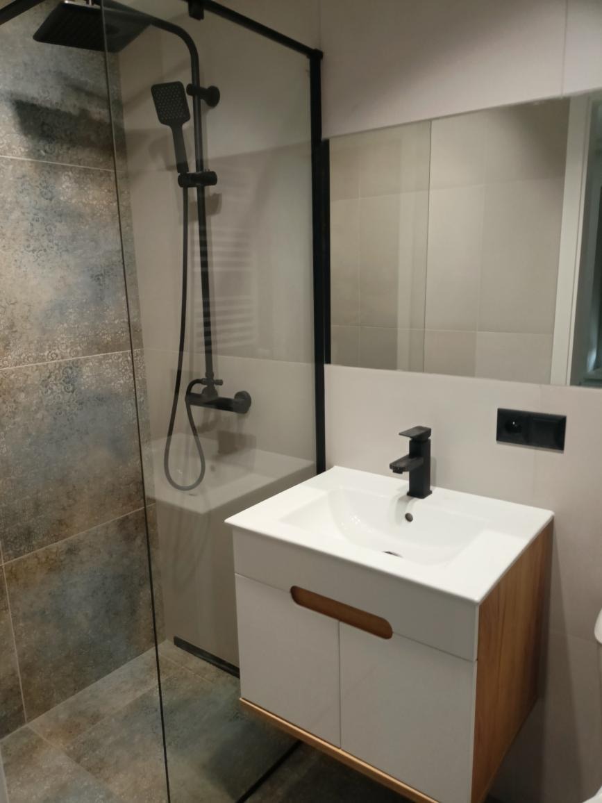 Nowoczesna łazienka z kabiną prysznicową typu walk-in z czarną armaturą, umywalką nablatową na szafce z drewnianymi akcentami i lustrem.