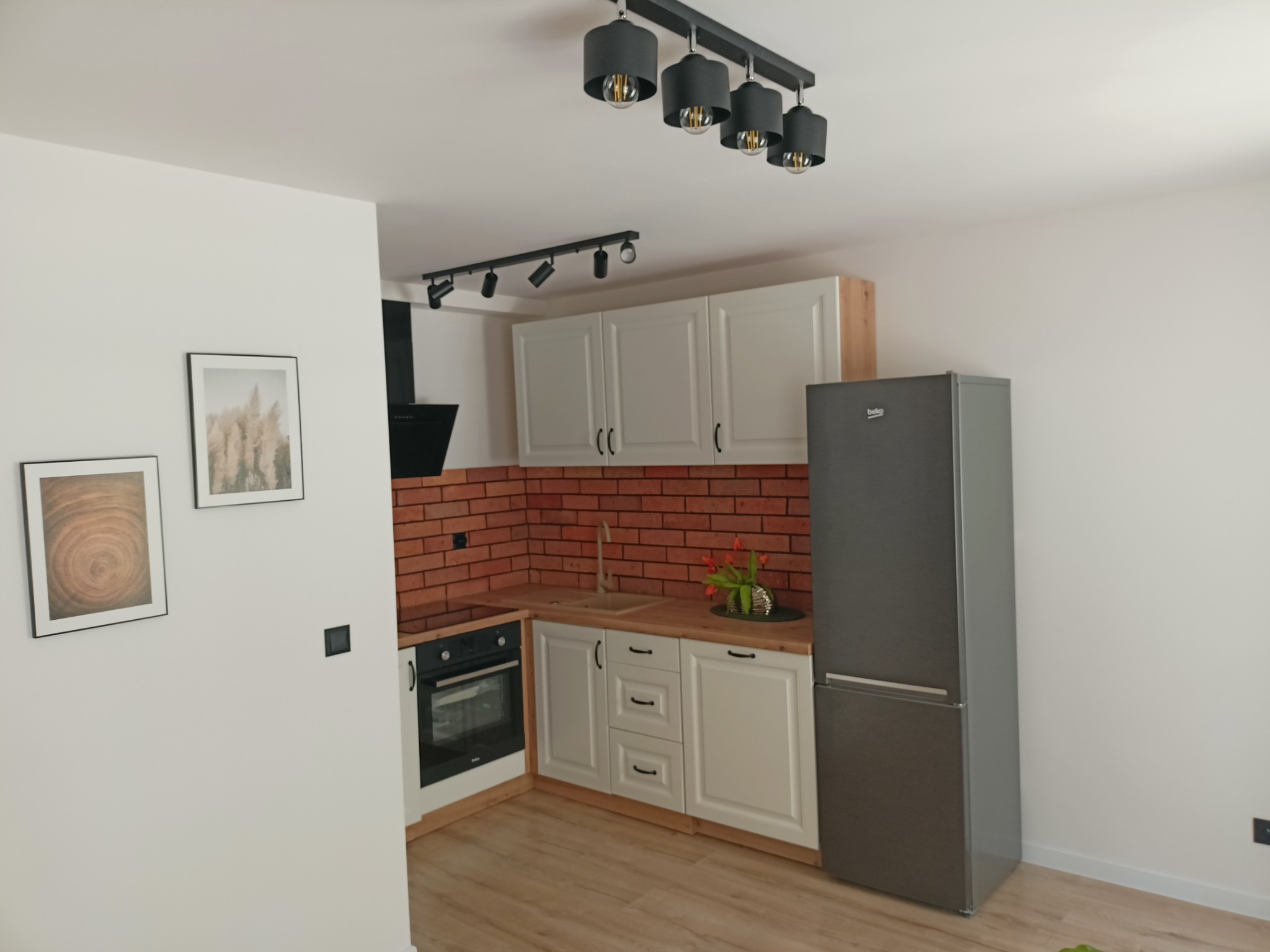 Nowoczesna kuchnia z białymi szafkami, drewnianym blatem, ceglaną ścianą nad blatem, szarą lodówką Beko i minimalistycznym oświetleniem sufitowym.