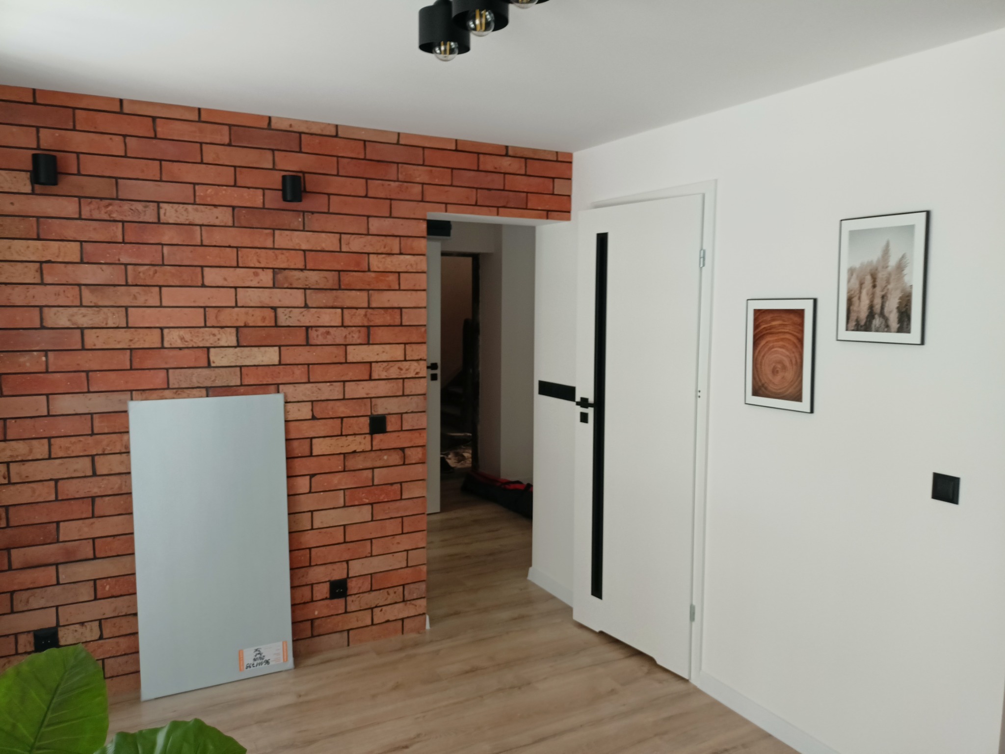 Ściana z cegły klinkierowej z czarnymi kinkietami, biała ściana z dwoma obrazami w czarnych ramkach, białe drzwi z czarnym paskiem, jasny parkiet i płyta gipsowa oparta o ścianę.