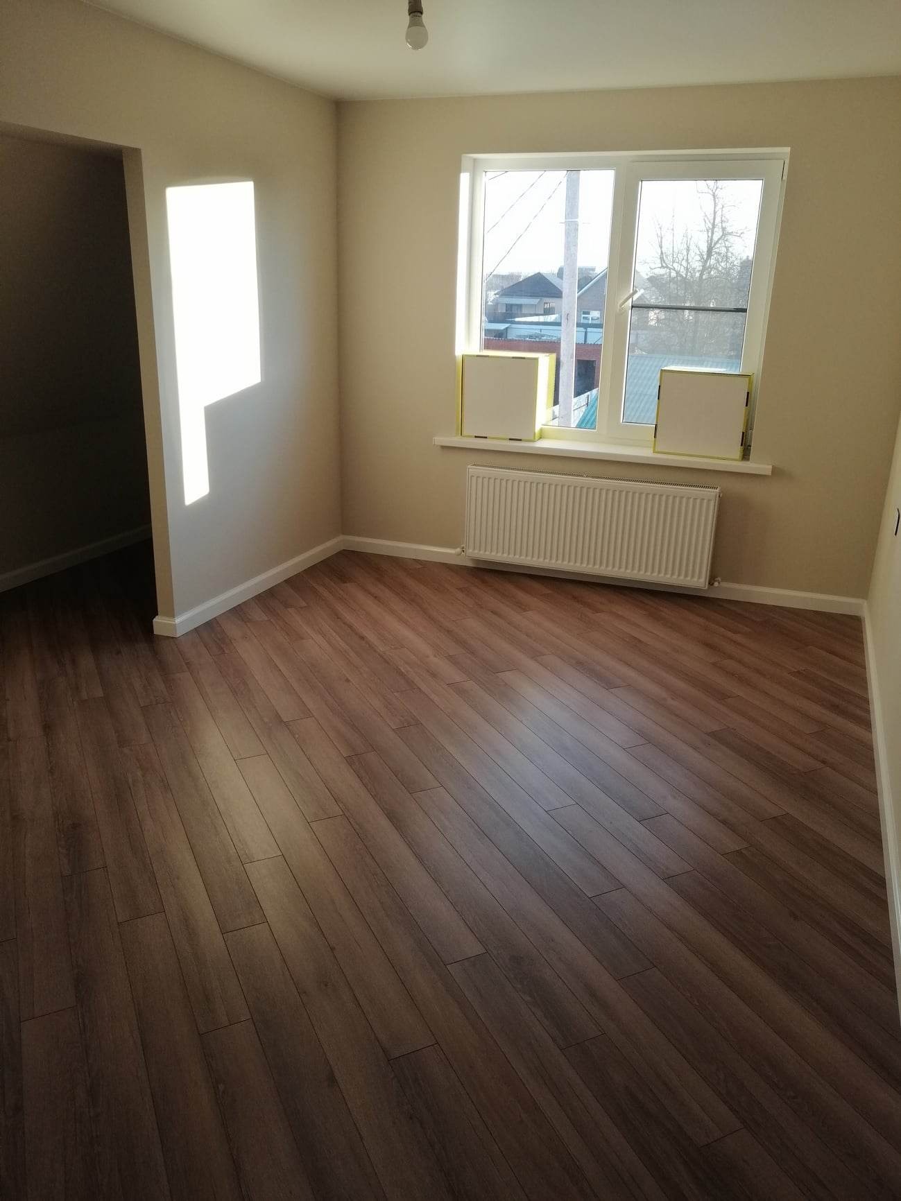 Jasne, puste pomieszczenie z panelami podłogowymi w odcieniach brązu, białymi ścianami i grzejnikiem pod oknem, z widocznymi kartonami na parapecie.