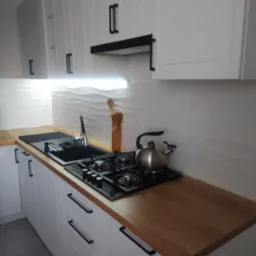 Nowoczesna kuchnia z białymi szafkami z czarnymi uchwytami, drewnianym blatem, czarnym zlewem i płytą gazową, oraz czajnikiem ze stali nierdzewnej na płycie.