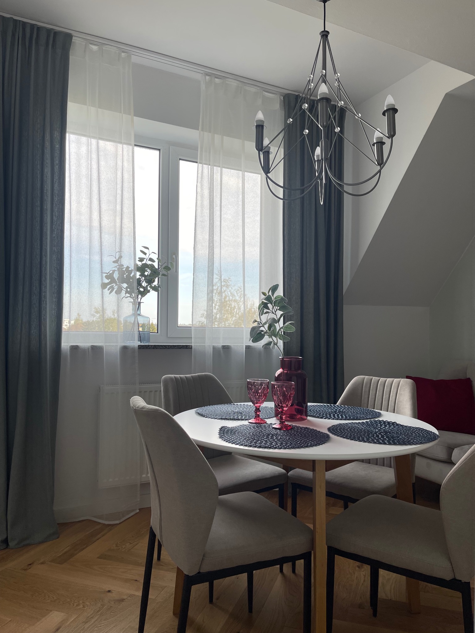 Jasne wnętrze jadalni z okrągłym stołem, szarymi krzesłami, dekoracjami i oknem z firanami. Nad stołem wisi nowoczesny żyrandol.