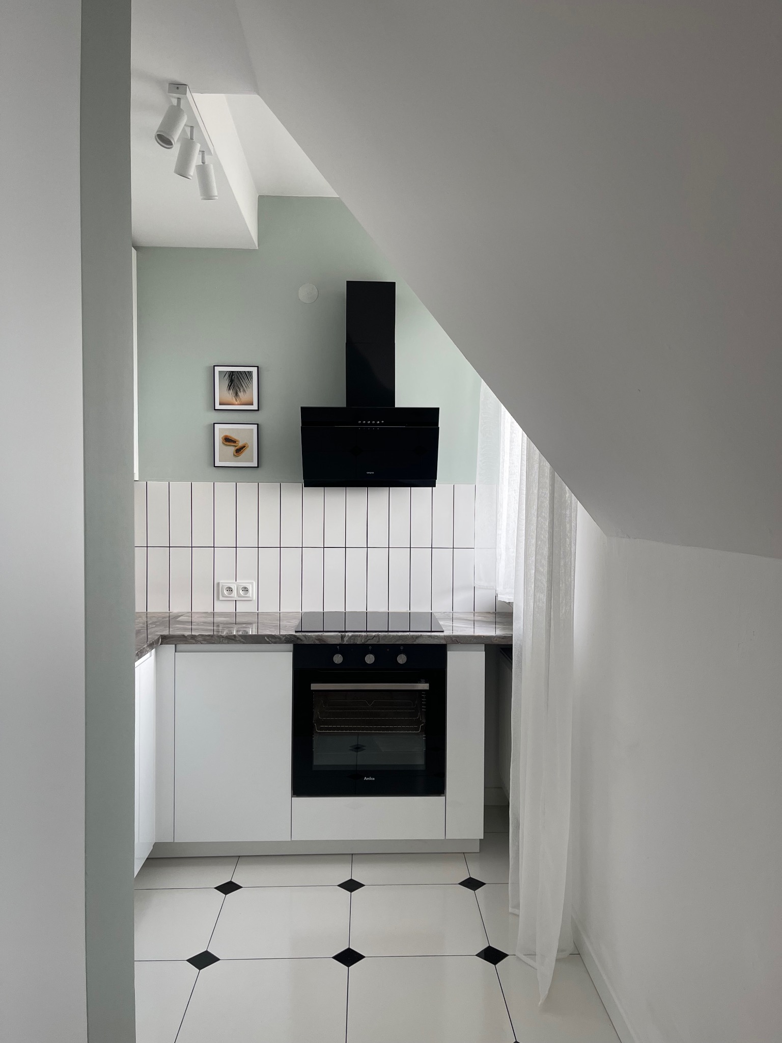 Jasna kuchnia w skandynawskim stylu z czarnymi akcentami: okap, piekarnik, płytki. Uwagę zwracają dekoracje ścienne i nietypowe oświetlenie.