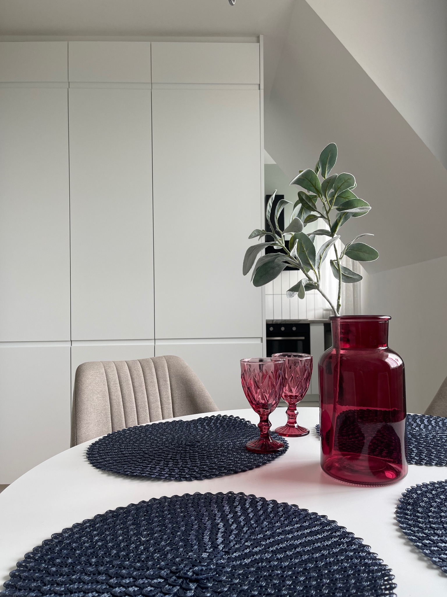 Biały stół z dekoracjami: czerwony wazon, kieliszki i podkładki. W tle zabudowa kuchenna i krzesło. Aranżacja wnętrza w jasnych barwach, minimalistyczny styl.