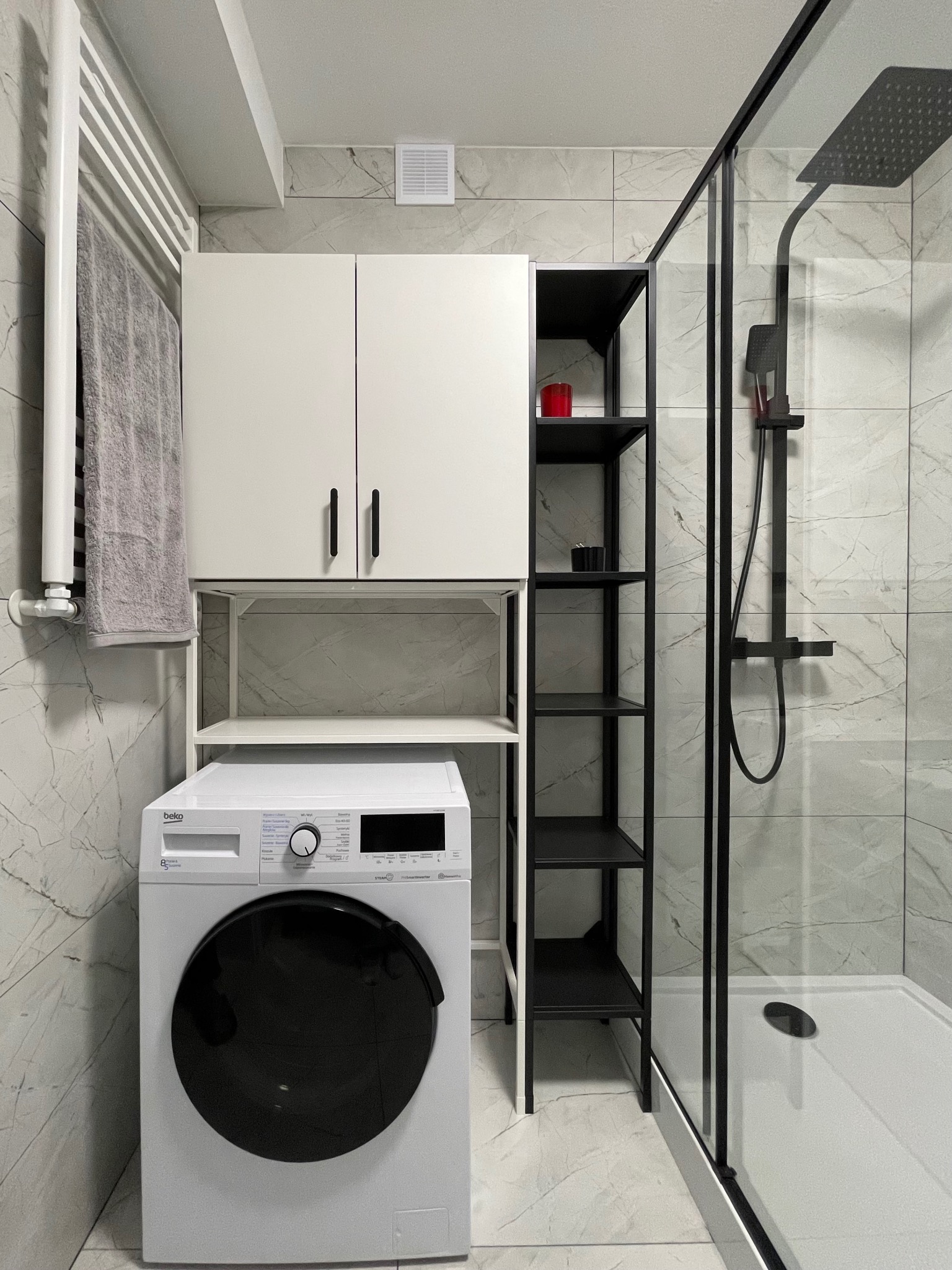 Nowoczesna łazienka w bieli i czerni: pralka, szafka nad nią, czarny regał i kabina prysznicowa z deszczownicą na tle marmurowych płytek.