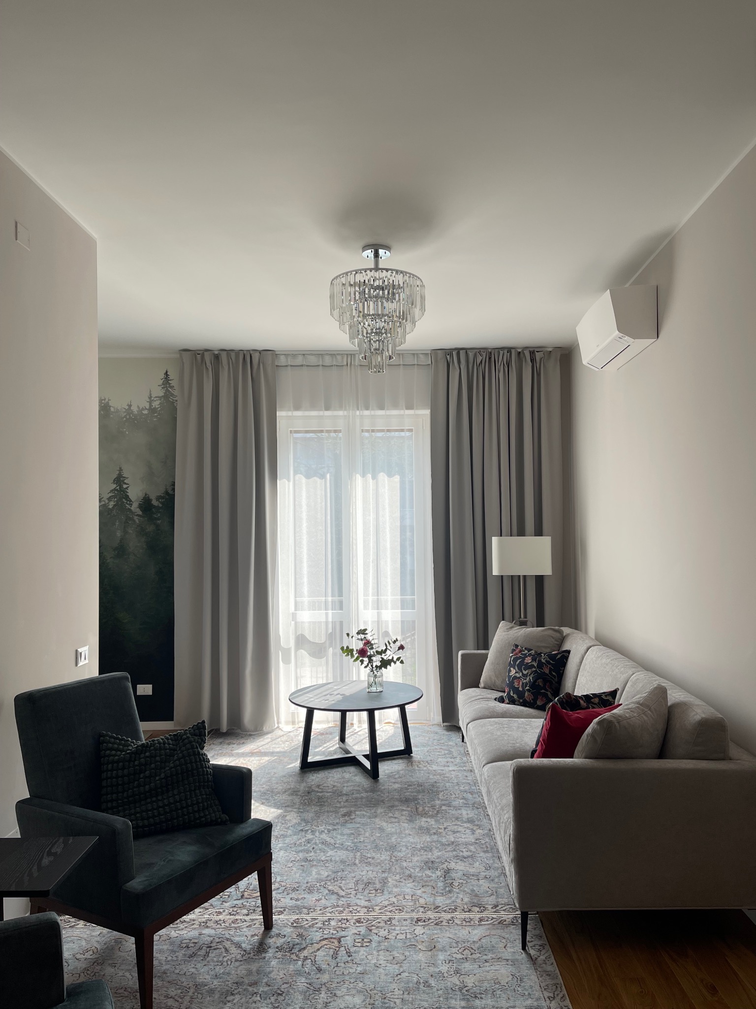 Jasny salon z szarą sofą, fotelami i okrągłym stolikiem kawowym. Ściana zdobiona tapetą z motywem leśnym. Kryształowy żyrandol dodaje elegancji.