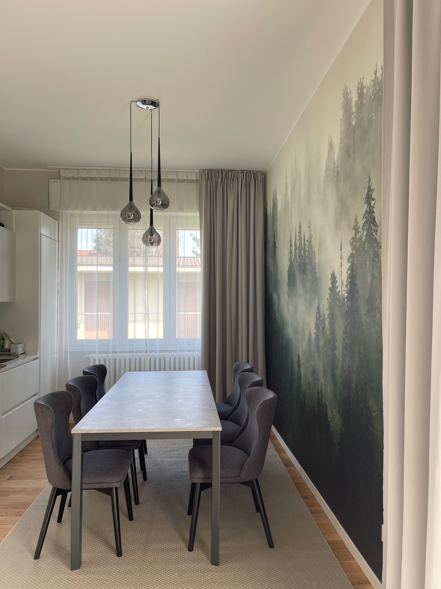 Jadalnia z tapetą leśną i szarymi krzesłami. Nowoczesny wystrój, wiszące lampy. Jasne wnętrze z akcentem natury, dywan w neutralnym odcieniu.