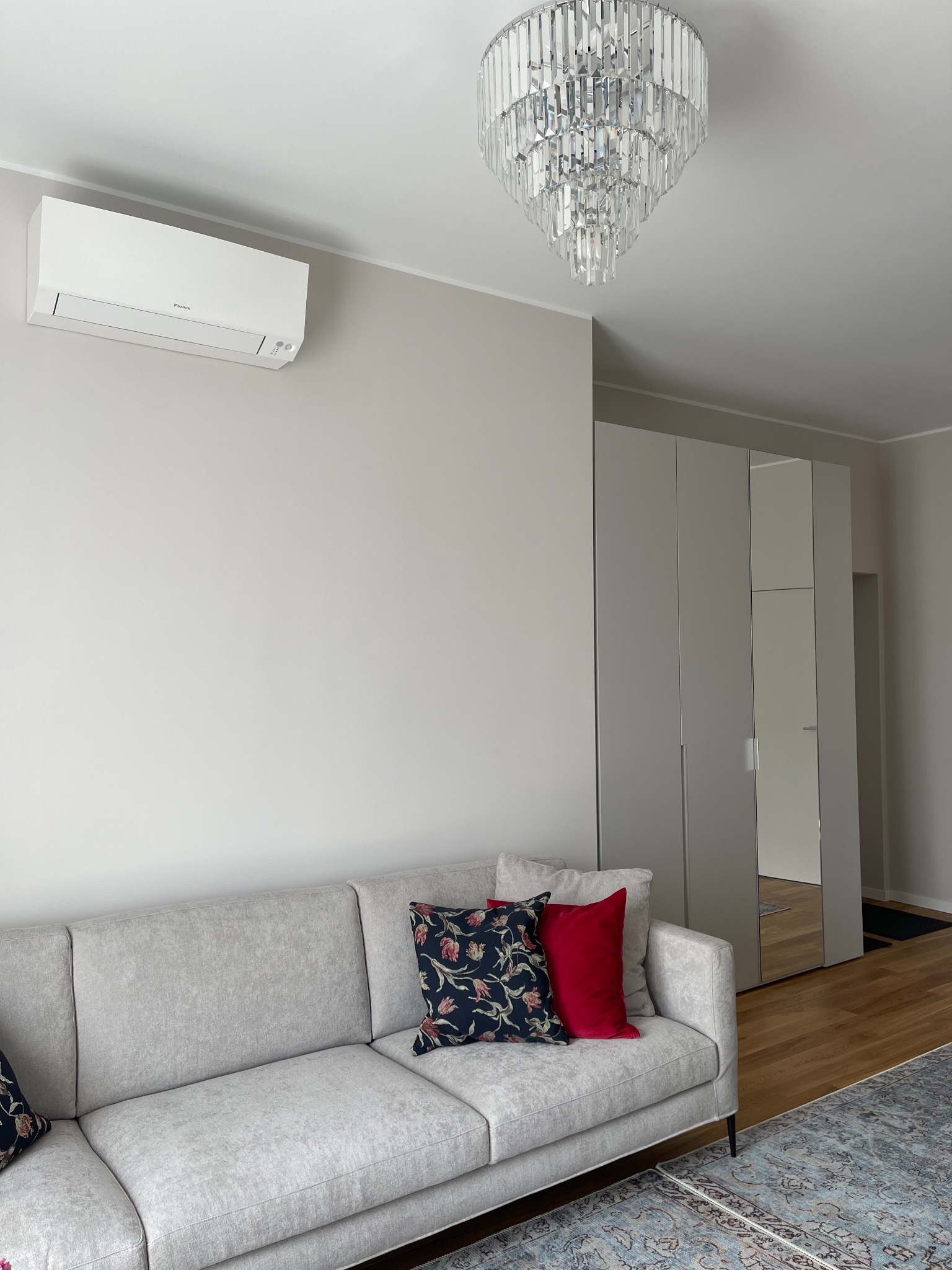 Jasny salon z szarą sofą, wzorzystymi poduszkami i kryształowym żyrandolem. Na ścianie klimatyzator, obok szafa z lustrami. Dywan z orientalnym wzorem.