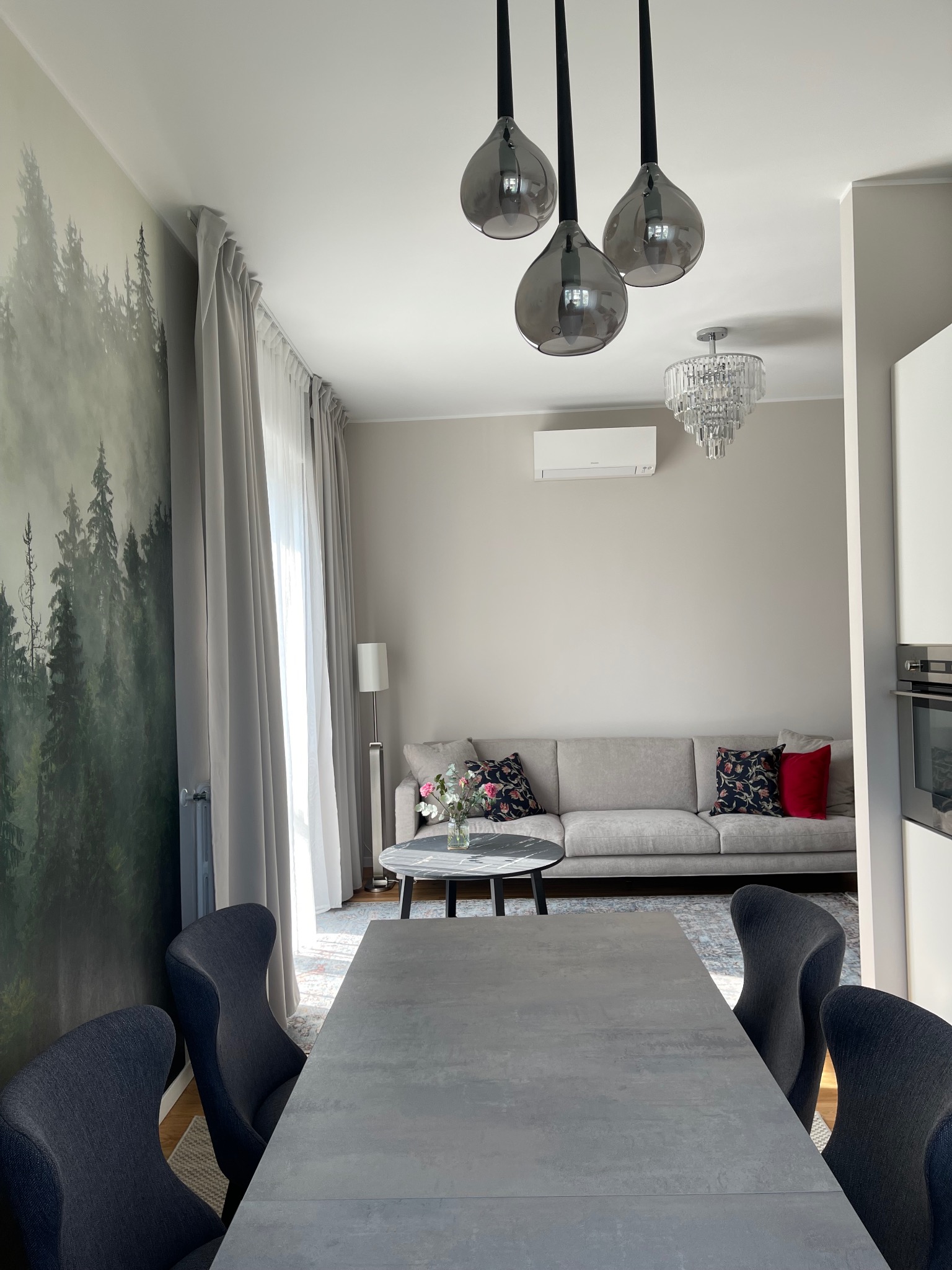 Nowoczesne wnętrze: szary stół z krzesłami, sofa, lampy wiszące i fototapeta z motywem lasu tworzą spójną, stylową przestrzeń w neutralnych barwach.