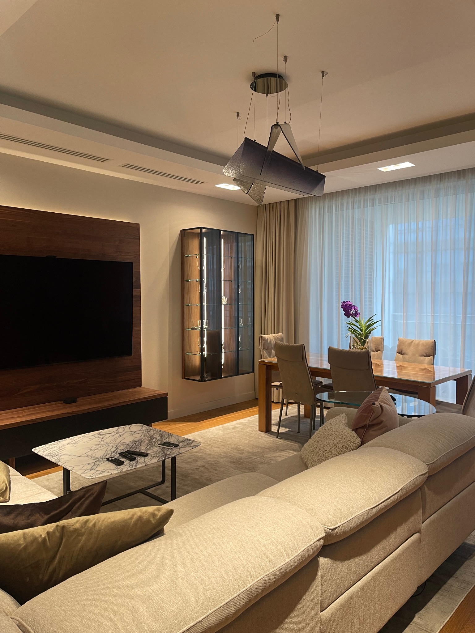 Salon z narożnikiem w beżowej tonacji, marmurowym stolikiem i drewnianą ścianą TV. Nad stołem jadalnym designerska lampa. Całość w stonowanej kolorystyce.