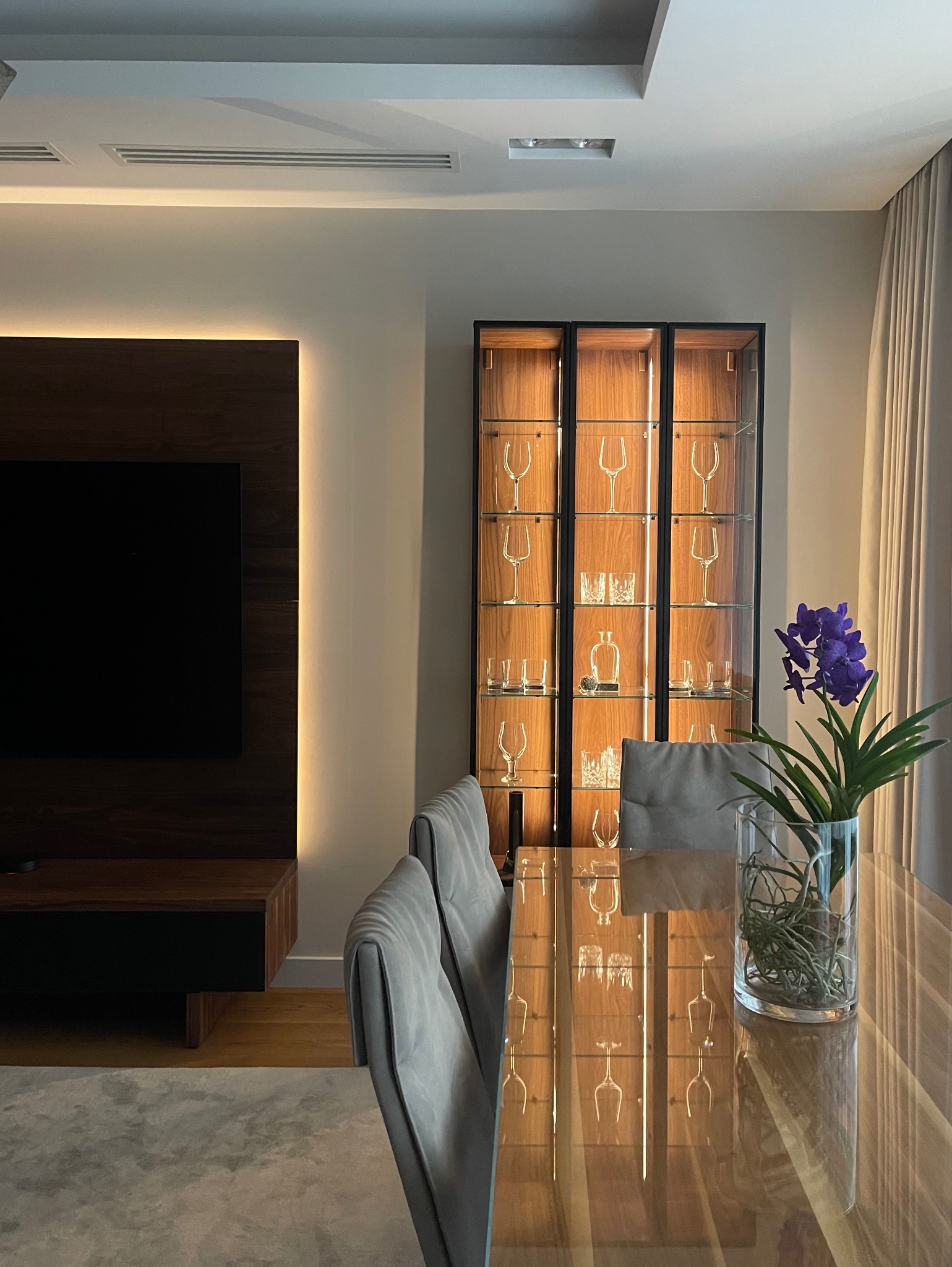 Elegancka jadalnia z podświetlaną witryną, szklanym stołem i kwiatami. Minimalistyczny design wnętrza z drewnianymi akcentami i subtelnym oświetleniem.