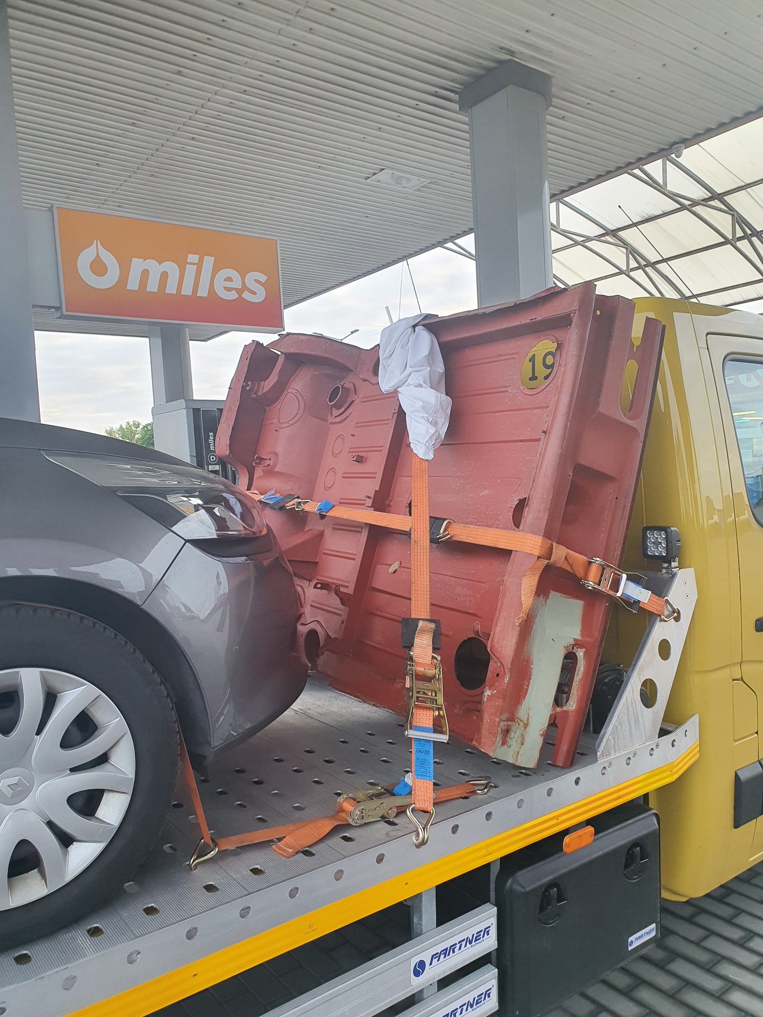 Żółta platforma transportowa z pomarańczowymi pasami mocującymi przewozi element karoserii samochodu osobowego w kolorze czerwonym z numerem 19 na tle stacji benzynowej.