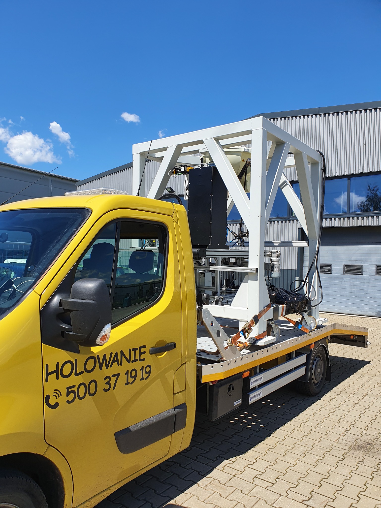 Żółty samochód z napisem 'HOLOWANIE' przewozi na platformie ładunek o konstrukcji stalowej, prawdopodobnie element maszyny przemysłowej, zabezpieczony pasami transportowymi.