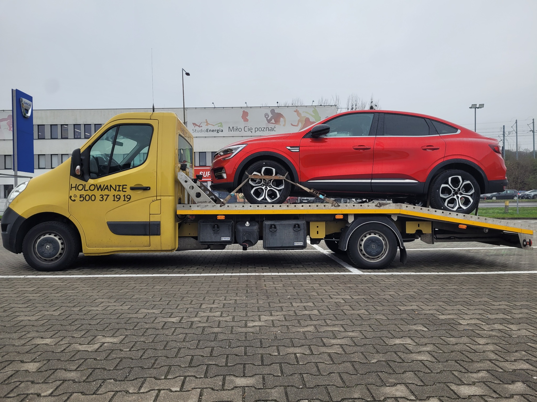 Żółty samochód pomocy drogowej z czerwonym samochodem na platformie, stojący na parkingu z kostki brukowej, w tle budynek z logo Dacia i billboardem.