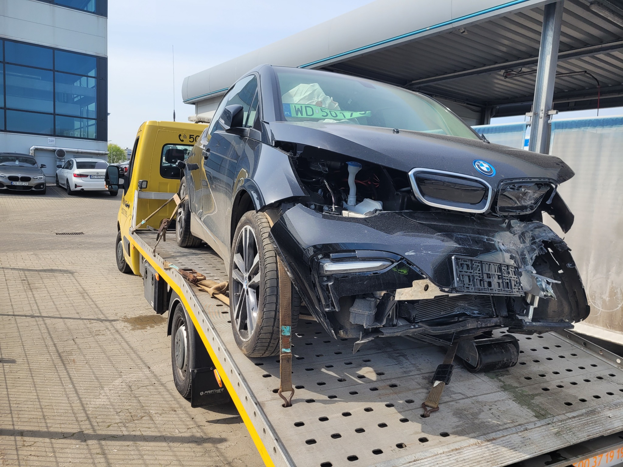 Uszkodzony, czarny samochód BMW i3 zabezpieczony pasami transportowymi na żółtej lawecie, widok z przodu na tle budynku z szklaną fasadą.