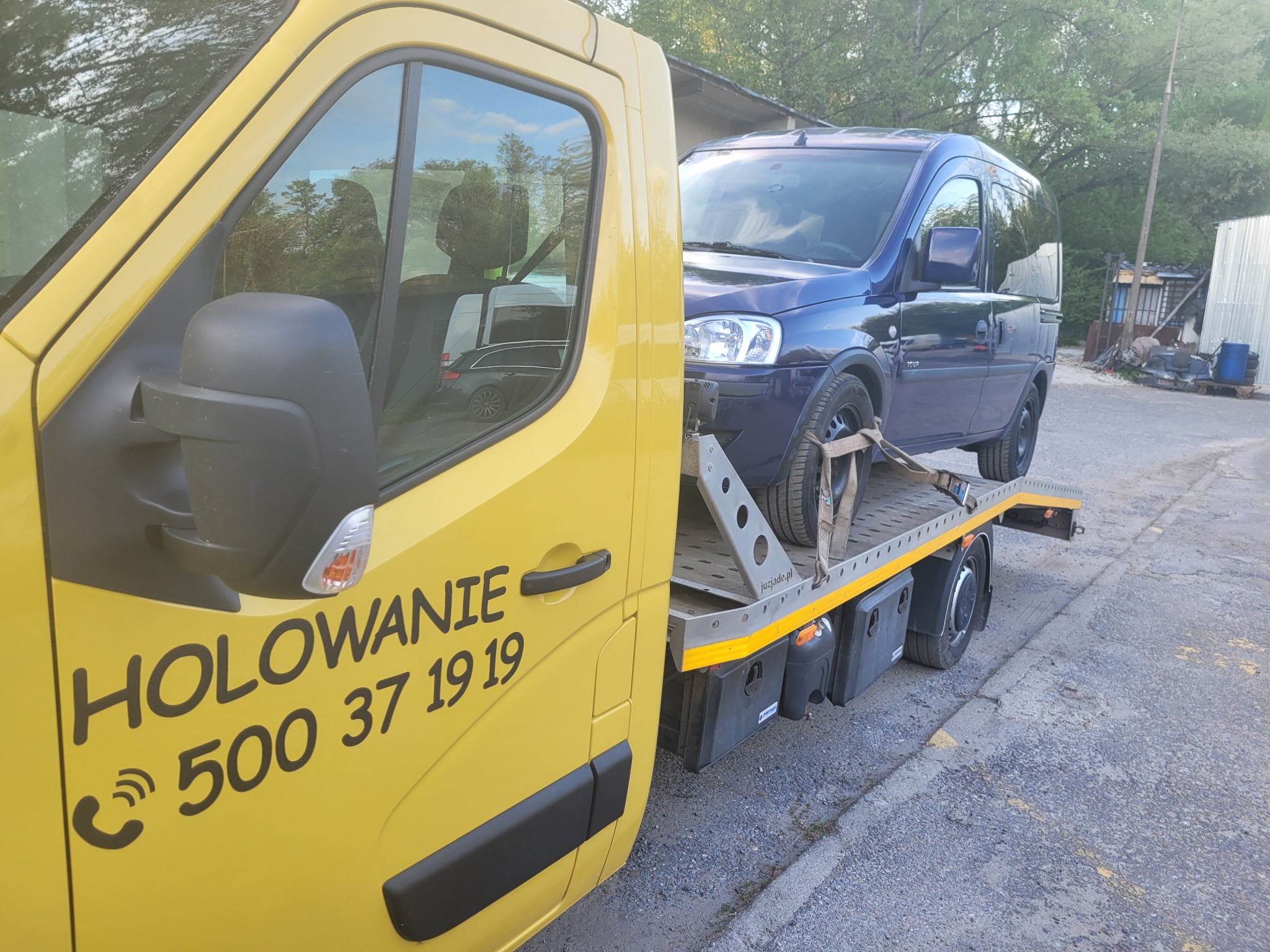 Żółta ciężarówka pomocy drogowej z numerem telefonu, przewożąca na platformie niebieski samochód osobowy.
