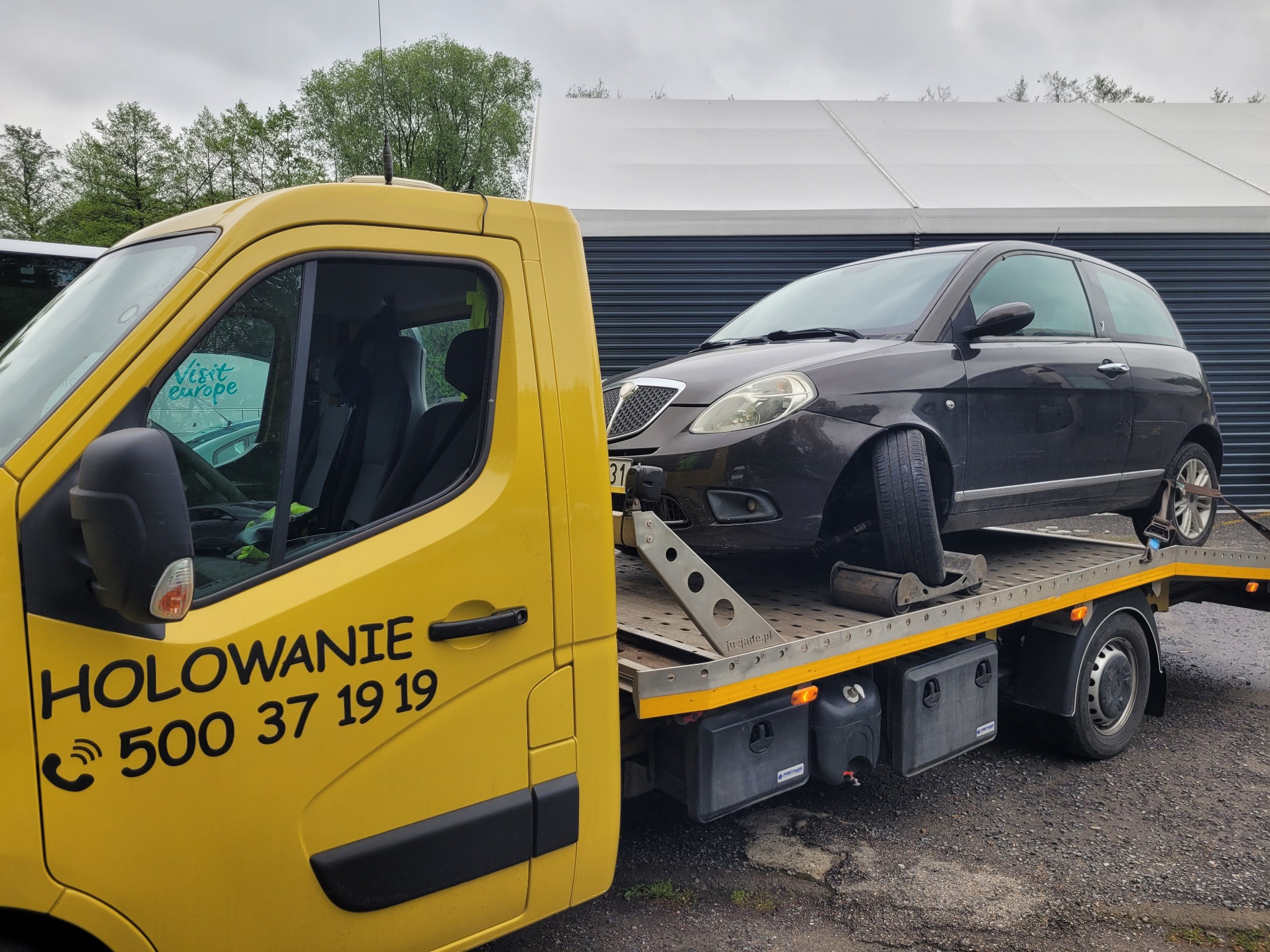 Żółty pojazd pomocy drogowej z napisem 'HOLOWANIE' i numerem telefonu, transportujący uszkodzony czarny samochód osobowy na platformie.