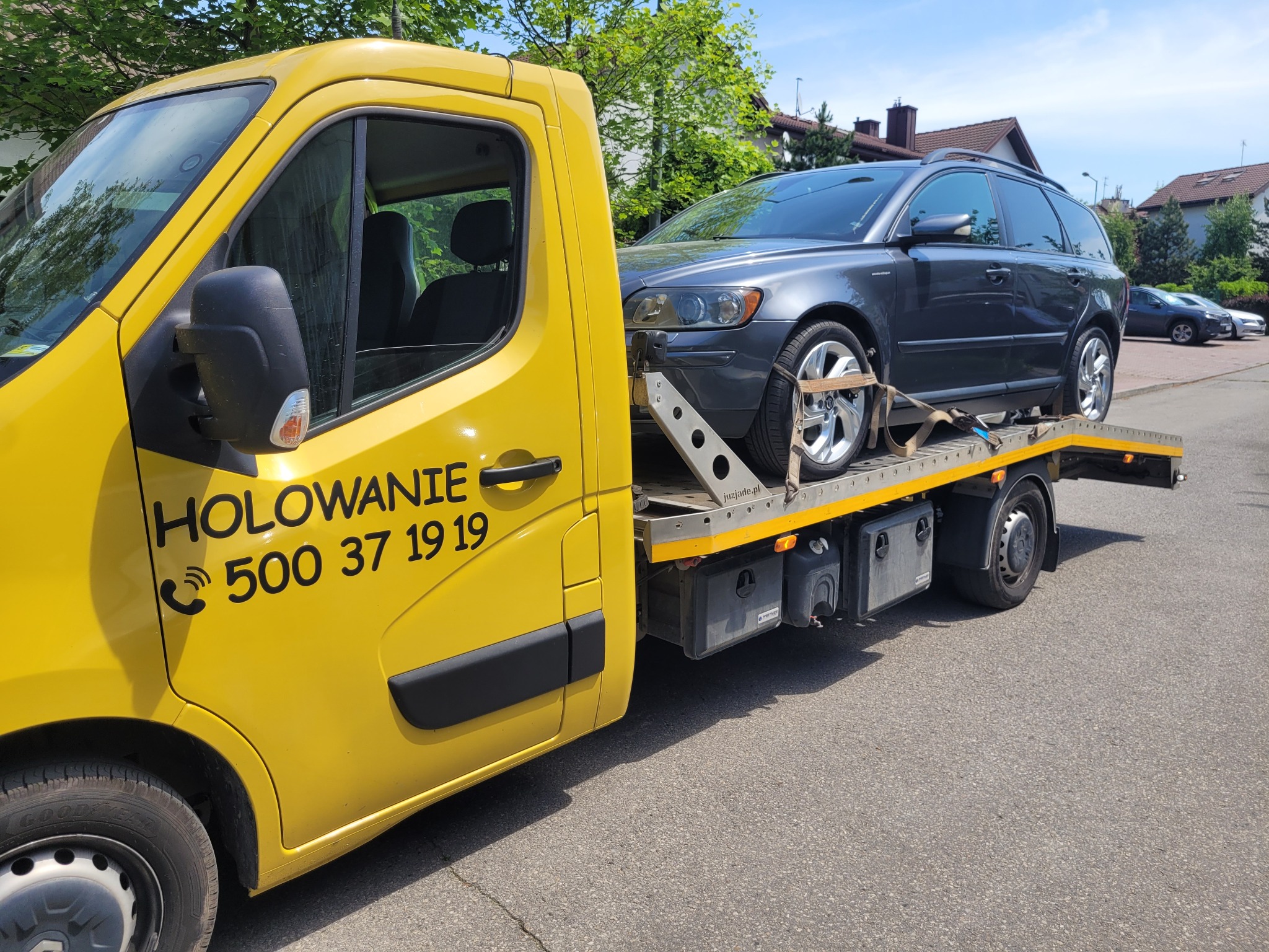 Żółta ciężarówka z numerem telefonu 'HOLOWANIE 500 37 19 19' przewozi szare kombi Volvo na platformie. Samochód zabezpieczony pasami transportowymi. Widoczne budynki mieszkalne w tle.