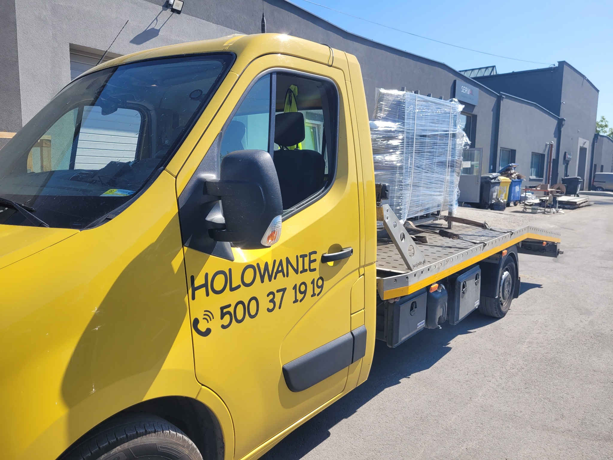 Żółty pojazd z napisem 'HOLOWANIE' i numerem telefonu przewozi zabezpieczony folią ładunek na platformie, zaparkowany na zewnątrz budynku firmy.