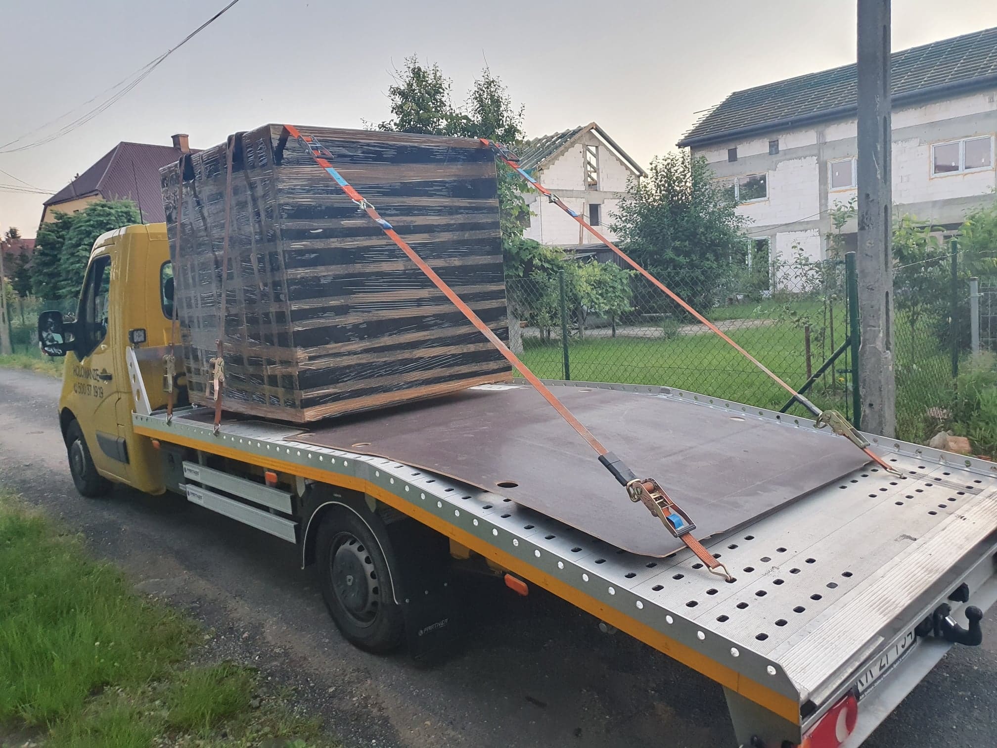 Żółta laweta z ładunkiem płyt drewnianych zabezpieczonych pomarańczowymi pasami transportowymi, na tle budynków mieszkalnych.