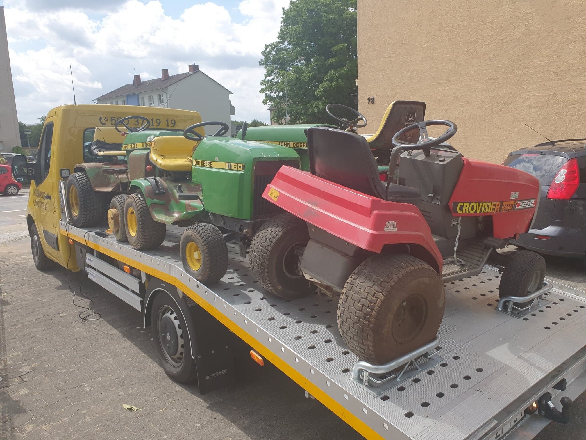 Żółta platforma transportowa z trzema różnymi traktorkami do koszenia trawy: zielony John Deere 160, czerwony Crovisier SBC550 i mniejszy, zielono-żółty model, zaparkowana na tle budynków...