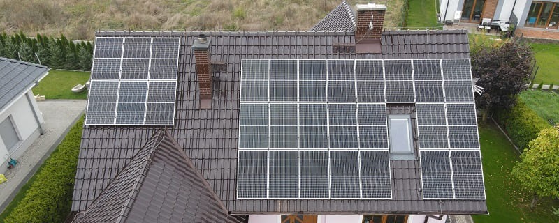 Instalacja fotowoltaiczna na dachu domu z brązową dachówką, widok z góry, panele fotowoltaiczne rozmieszczone symetrycznie wokół okna dachowego i komina.
