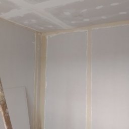 Prywatna - Pokój w trakcie montażu płyt kartonowo-gipsowych na ścianach i suficie, widoczne łączenia płyt zabezpieczone taśmą, drabina oparta o ścianę, surowy stan wykończenia.