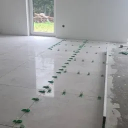 Układanie jasnych płytek podłogowych z użyciem zielonych klipsów poziomujących i sznurka w nowym, jasnym pomieszczeniu z dużym oknem.