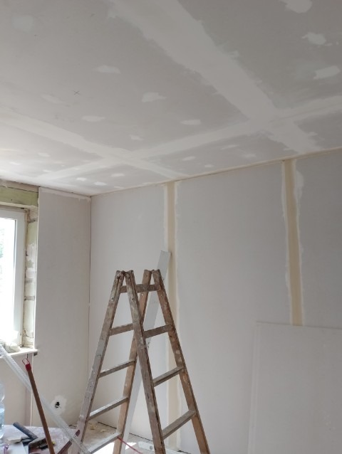 Montaż płyt karton-gips na ścianach i suficie w trakcie szpachlowania, widoczna drewniana drabina i narzędzia.