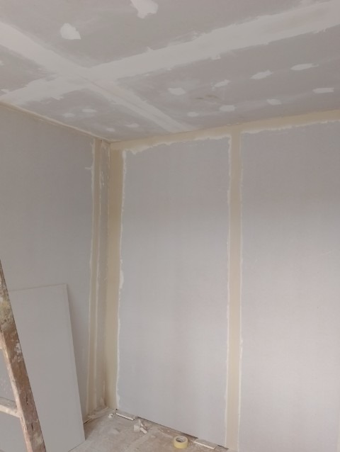 Pokój w trakcie montażu płyt kartonowo-gipsowych na ścianach i suficie, widoczne łączenia płyt zabezpieczone taśmą, drabina oparta o ścianę, surowy stan wykończenia.